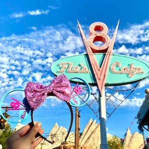 May include: Pink and blue Minnie Mouse ears with a pink bow and a blue and pink "Flo's Cafe" sign. The sign is in front of a blue sky with white clouds.