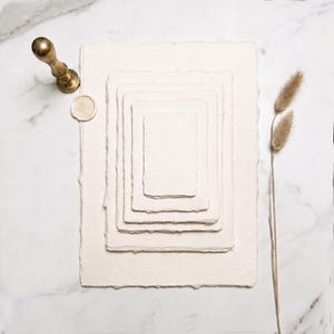 Peut inclure: Une pile de cinq feuilles rectangulaires de papier fait main blanc cassé aux bords effilochés, disposées par taille décroissante. Un sceau en laiton et un cachet de cire circulaire sont placés à gauche, avec deux tiges d'herbe séchée à droite.