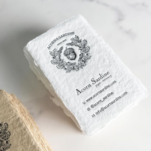 May include: A stack of white paper stationery with a deckled edge. The paper has a printed logo of an acorn and fish inside a wreath. The text "Acorn & Sardine" and contact information are printed on the paper.