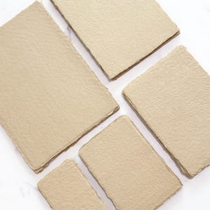 May include: A collection of handmade, beige-colored paper sheets with rough, textured surfaces and deckled edges. The sheets are arranged on a white background, showcasing their natural, organic appearance. The paper appears to be suitable for writing, drawing, or crafting.
