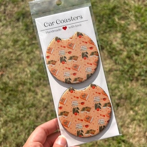May include: Two car coasters with a repeating pattern of books, coffee cups, and apples on a peach background. The coasters are packaged in a clear plastic sleeve with the text "Car Coasters" and "Handmade with love".
