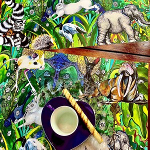 May include: Two placemats with a colorful jungle animal print design. The placemats feature a variety of animals, including elephants, mice, birds, and a rabbit. A purple heart-shaped saucer with a white teacup and a long, thin, brown cookie sits on top of the placemats.