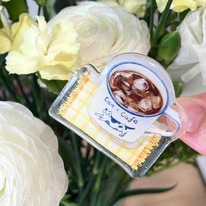 May include: A clear acrylic pin featuring a white mug filled with iced coffee. The mug has a blue rim and the words "Cat Cafe" with a cat illustration. The pin rests on a yellow checkered background.