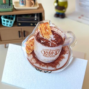 May include: A clear acrylic standee featuring a detailed illustration of a white coffee mug filled with dark brown liquid and a golden-brown waffle. The mug displays the text Little-drumsticks 1059-3 Coffee House. The saucer is covered in brown powder.