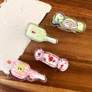 May include: Four clear acrylic charms with colorful designs. One charm is a green bottle with a rose, another is a pink bottle with a floral design, and two are candy-shaped with strawberry and lemon designs. The charms are on a wooden surface.