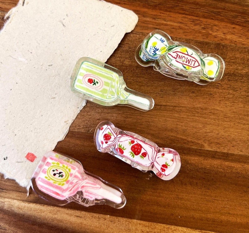 May include: Four clear acrylic charms with colorful designs. One charm is a green bottle with a rose, another is a pink bottle with a floral design, and two are candy-shaped with strawberry and lemon designs. The charms are on a wooden surface.