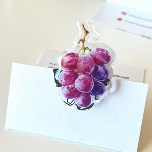 May include: A clear acrylic pin featuring a detailed illustration of a cluster of purple and red grapes with a brown stem. The pin is attached to a white rectangular card that displays printed text, including the word lightweight.