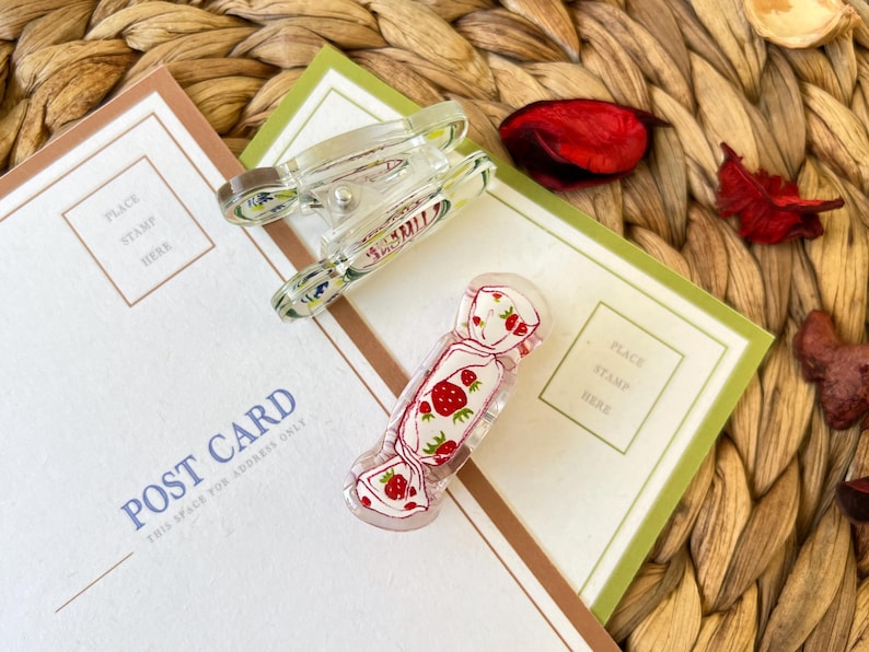 May include: Two clear acrylic hair clips and a candy-shaped clip with a strawberry design rest on a postcard. The clips are decorated with red and green strawberries on a white background. The postcard has a place for a stamp.