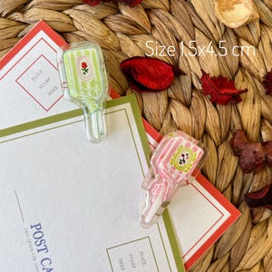May include: Two clear acrylic paper clips, one green and one pink, shaped like vintage perfume bottles, each with a floral design. The clips are 15 cm x 4.5 cm and rest on postcards with red and green borders.
