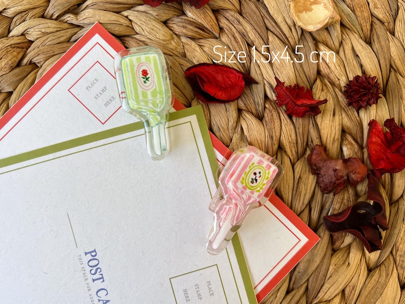May include: Two clear acrylic paper clips, one green and one pink, shaped like vintage perfume bottles, each with a floral design. The clips are 15 cm x 4.5 cm and rest on postcards with red and green borders.