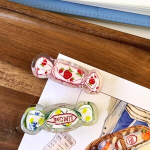 May include: Two clear acrylic hair clips shaped like wrapped candies. One clip features a strawberry design with red strawberries and green leaves. The other clip has the word "LIMONE" and a lemon design.