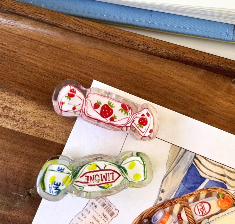 May include: Two clear acrylic hair clips shaped like wrapped candies. One clip features a strawberry design with red strawberries and green leaves. The other clip has the word "LIMONE" and a lemon design.