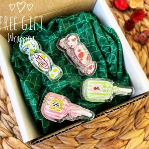 May include: A gift box with four clear acrylic candy-shaped charms. The charms include a strawberry, a lemon, and a bottle design. The box is lined with green patterned tissue paper. The text "FREE GIFT Wrapping" is visible on the box.