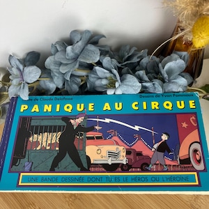May include: A vintage comic book titled "Panique au Cirque" with illustrations of a circus scene. The cover features a man in a suit and a boy, with text in yellow and blue. The book is on a wooden surface.