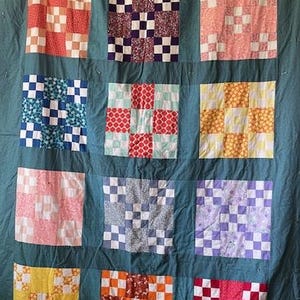 Może przedstawiać: Ręcznie robiona kołdra z tłem w kolorze morskim i dwunastoma kwadratowymi blokami. Każdy blok ma inny wzór patchworkowy w różnych kolorach, w tym czerwonym, różowym, fioletowym, niebieskim i żółtym. Kołdra wisi na drążku.