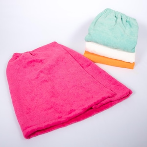 May include: A hot pink terry cloth hair wrap is displayed next to a stack of three similar wraps in mint green, white, and orange. The wraps appear soft and absorbent, ideal for post-shower use.