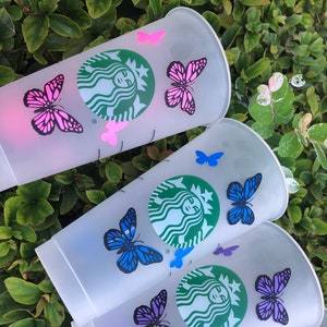 May include: Clear plastic Starbucks cold cups decorated with butterfly decals in various colours. The cups display the Starbucks logo in green and are adorned with pink, blue, and purple butterfly designs. The cups are set against a backdrop of green foliage.