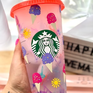 May include: A pink and white reusable cup with a Starbucks logo and ice cream cone designs. The cup has a red lid and a clear straw.
