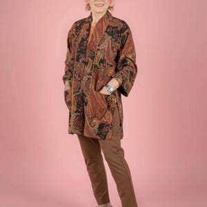 May include: A woman wearing a paisley print kimono-style jacket with brown pants and purple shoes. The jacket features a black background with red, brown, and gold paisley patterns. The outfit is set against a pink backdrop.