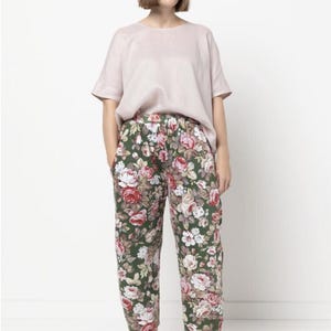 May include: A woman wearing a light pink short-sleeved top and floral print pants. The pants feature a dark green background with a pattern of red, pink, and white roses. She is also wearing pink sneakers.