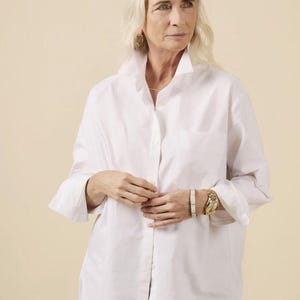 May include: A white button-down shirt with a classic collar and long sleeves. The shirt is worn with blue jeans. The model is wearing gold hoop earrings, a gold watch, and a white bracelet.