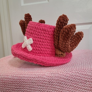May include: A pink crocheted hat with brown crocheted antlers and a white cross on the front.