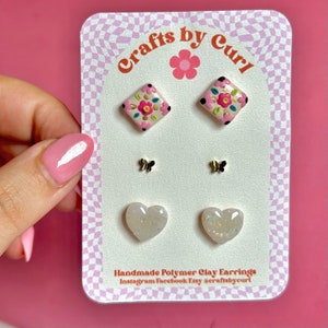 Op de afbeelding: Set handgemaakte polymer clay oorbellen met een paar roze en witte vierkante bloemen oorbellen, een paar witte hartvormige oorbellen met glitter en een paar gouden vlinder oorbellen. De oorbellen worden gepresenteerd op een witte kaart met de tekst "Crafts by Curl" en "Handmade Polymer Clay Earrings Instagram Facebook Etsy craftsbycurl".