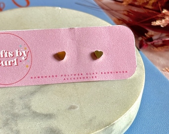 Pode incluir: Um par de brincos dourados pequenos em forma de coração em um cartão rosa. O cartão diz "Gifts by Curl" e "Handmade Polymer Clay Earrings & Accessories". Os brincos são exibidos em uma superfície redonda de pedra cinza claro.