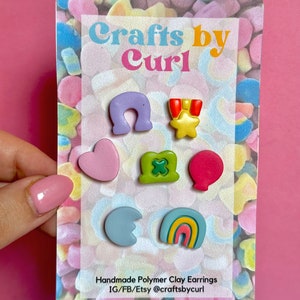 Op de afbeelding: Een set handgemaakte polymer clay oorbellen met verschillende vormen en kleuren, waaronder een hoefijzer, een ster met een lint, een hart, een klavertjevier, een ballon, een halve maan en een regenboog. De oorbellen worden gepresenteerd op een kaart met de tekst "Crafts by Curl" en "Handmade Polymer Clay Earrings IG/FB/Etsy @craftsbycurl".