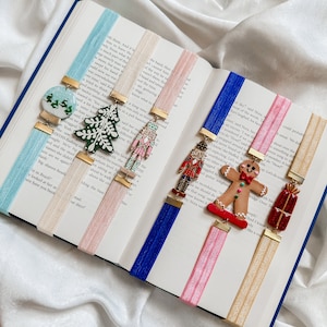 May include: A collection of eight Christmas-themed bookmarks with various designs, including a gingerbread man, a nutcracker, a Christmas tree, a gift box, and a snow globe. The bookmarks are made of metal and have a gold finish.