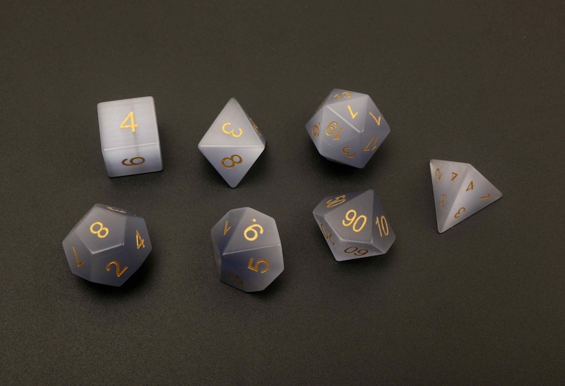 May include: Set of seven polyhedral dice in various shapes, including a cube, pyramid, and d20. The dice are a translucent gray color with gold-colored numbers. Ideal for tabletop games.
