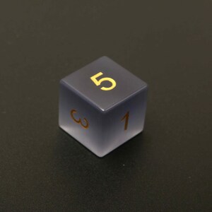 May include: A six-sided die with a gradient gray and frosted finish. The number 5 is prominently displayed in gold on the top face, with the numbers 1 and 3 visible on adjacent sides. A gaming accessory.