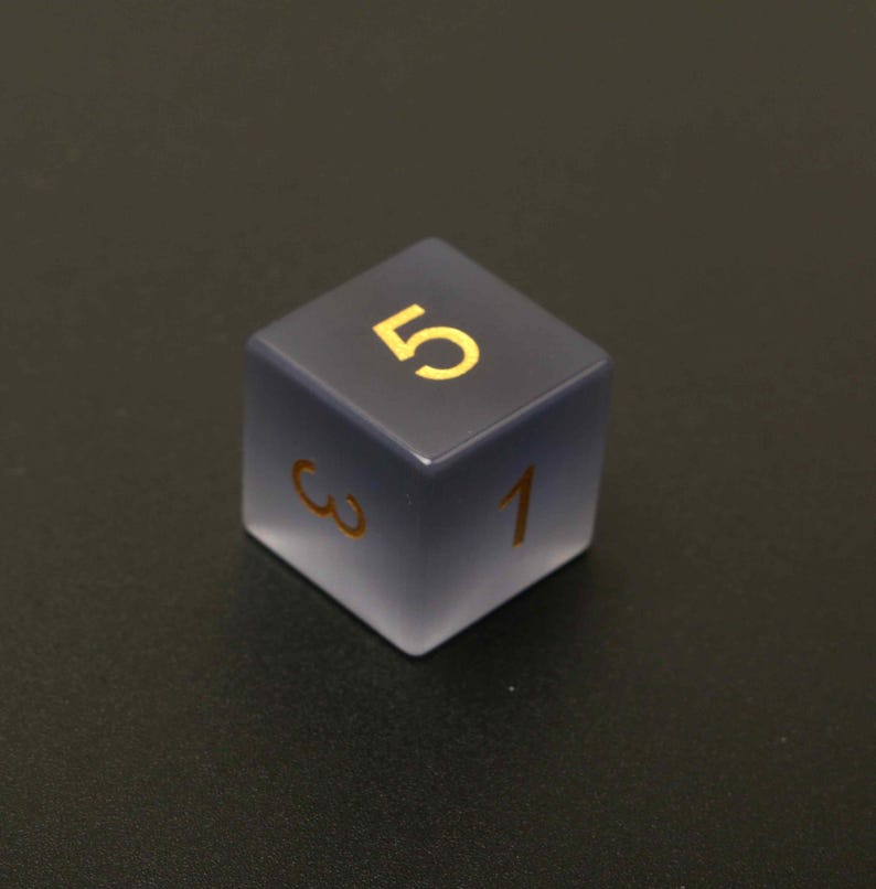 May include: A six-sided die with a gradient gray and frosted finish. The number 5 is prominently displayed in gold on the top face, with the numbers 1 and 3 visible on adjacent sides. A gaming accessory.