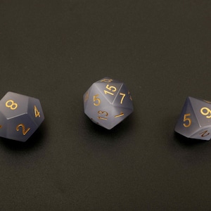 May include: Three polyhedral dice with gold-colored numbers. The dice are a smoky gray color and are arranged on a dark surface. The dice are used for tabletop games.