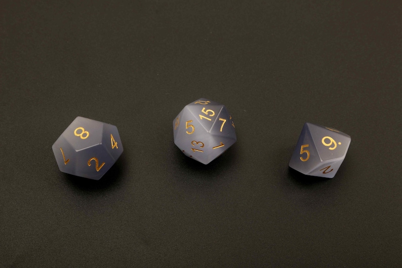 May include: Three polyhedral dice with gold-colored numbers. The dice are a smoky gray color and are arranged on a dark surface. The dice are used for tabletop games.