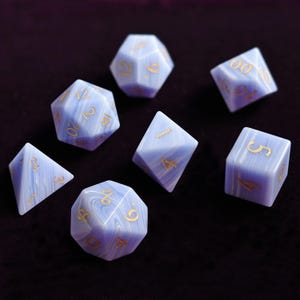 Peut inclure: Un ensemble de sept dés polyédriques bleus clairs avec un effet marbré, comprenant un d4, d6, d8, d10, d12, d20 et un dé de pourcentage. Les chiffres sont dorés. Les dés sont posés sur une surface violet foncé.