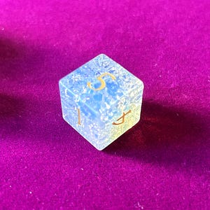 May include: A translucent, blue-toned, six-sided die with gold-colored numerals. The die is set against a deep purple background. The numbers 1, 4, and 5 are visible on the die's faces. This is a gaming accessory.