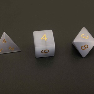 May include: Three dice for tabletop games. The set includes a four-sided die, a six-sided die, and an eight-sided die. Each die is a translucent gray color with gold-colored numbers. The dice are arranged on a dark gray surface.
