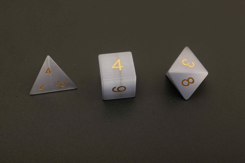 May include: Three dice for tabletop games. The set includes a four-sided die, a six-sided die, and an eight-sided die. Each die is a translucent gray color with gold-colored numbers. The dice are arranged on a dark gray surface.