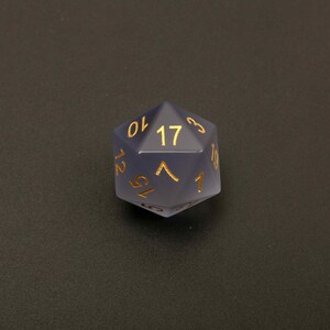 May include: A translucent, blue-grey, twenty-sided die with gold-colored numerals. The die is a polyhedron, with the number 17 prominently displayed on one face. The numbers are in a serif font. The die is designed for tabletop role-playing games.