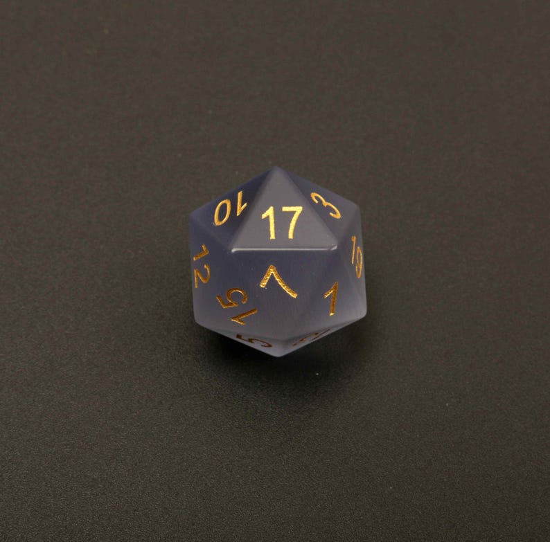 May include: A translucent, blue-grey, twenty-sided die with gold-colored numerals. The die is a polyhedron, with the number 17 prominently displayed on one face. The numbers are in a serif font. The die is designed for tabletop role-playing games.
