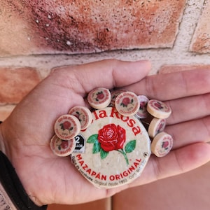 Op de afbeelding: Een hand die een aantal kleine ronde snoepjes vasthoudt met een grotere ronde snoepdoos in het midden. De doos is gelabeld "La Rosa Mazapan Original" met een rood rozenontwerp. De tekst op de doos luidt "Dulce de cacahuate. Original desde 1950. Cont. Net. 280g."