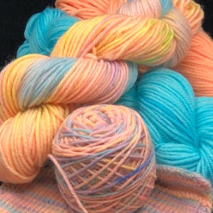 May include: A collection of colorful yarn skeins in shades of peach, yellow, blue, and pink. A ball of yarn and a small knitted swatch are also visible. The yarn appears soft and suitable for knitting or crochet projects.