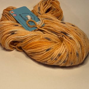 May include: A skein of hand-dyed yarn in shades of orange, yellow, and gray. The yarn is loosely wound, showing the texture of the fibers. A blue tag with the text "Artisan Hand Dyed" is attached to the yarn.