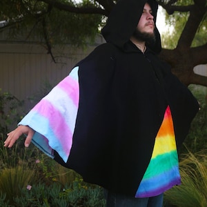May include: A black hooded cloak with tie-dye detailing. The sleeves have a pastel pink and blue tie-dye pattern, and the side panels feature a rainbow design. The cloak is worn outdoors.