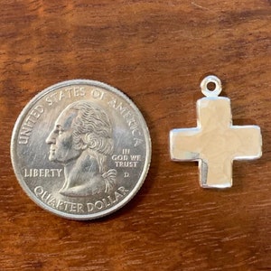 Pu&ograve; includere: Un ciondolo a forma di croce in argento, mostrato accanto a un quarto di dollaro americano per il confronto delle dimensioni. La croce &egrave; leggermente testurizzata e ha un design semplice.