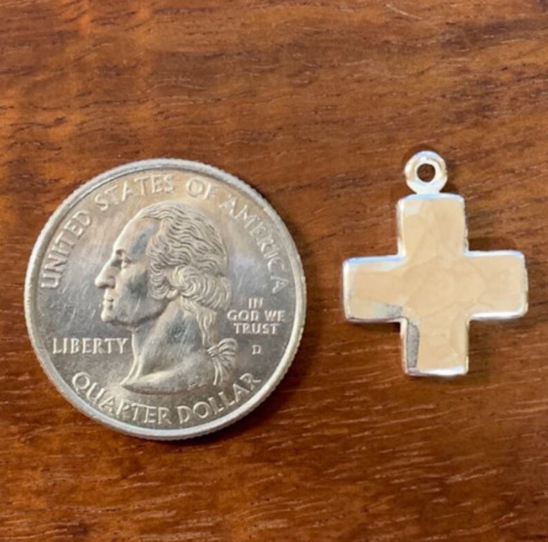 Pu&ograve; includere: Un ciondolo a forma di croce in argento, mostrato accanto a un quarto di dollaro americano per il confronto delle dimensioni. La croce &egrave; leggermente testurizzata e ha un design semplice.