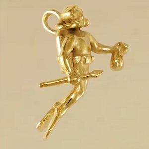 May include: A gold-colored charm in the shape of a diver, holding a spear and a pouch. The diver is wearing a diving helmet and gear. The charm has a loop at the top for attaching to a necklace or bracelet.