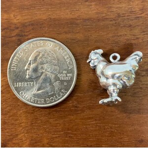 K&ouml;nnte beinhalten: Ein silberner Anh&auml;nger in Form eines Hahns, neben einem US-amerikanischen Viertel f&uuml;r den Gr&ouml;&szlig;envergleich. Der Anh&auml;nger hat eine Schlaufe zum Anbringen an einem Armband oder einer Halskette.