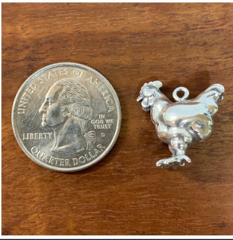 K&ouml;nnte beinhalten: Ein silberner Anh&auml;nger in Form eines Hahns, neben einem US-amerikanischen Viertel f&uuml;r den Gr&ouml;&szlig;envergleich. Der Anh&auml;nger hat eine Schlaufe zum Anbringen an einem Armband oder einer Halskette.